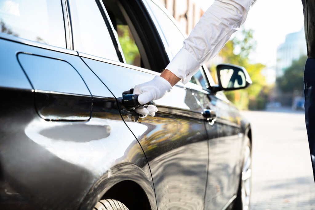 A chauffeur in a white dress shirt and gloves is opening the door of a black luxury car. The scene suggests service or arrival.